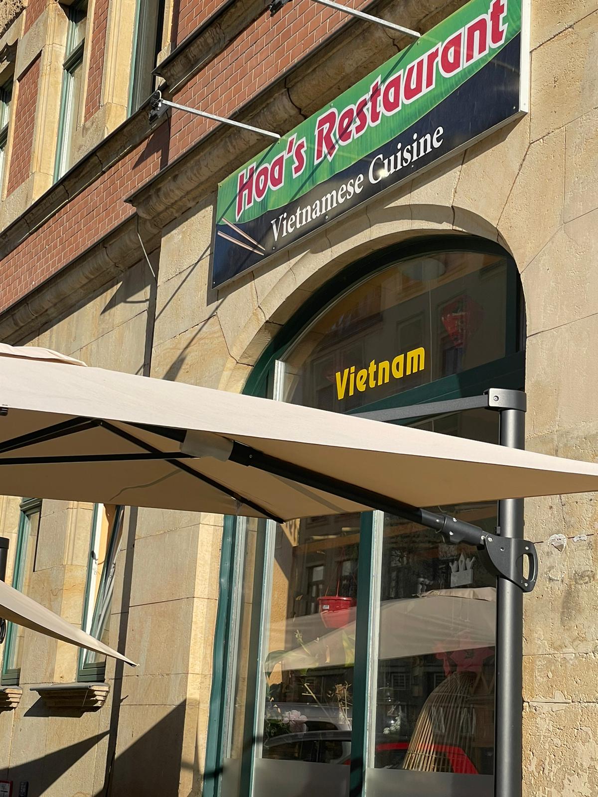 Außenansicht von Hoa's Restaurant mit Schildern und Sonnenschirm, warmes Tageslicht.