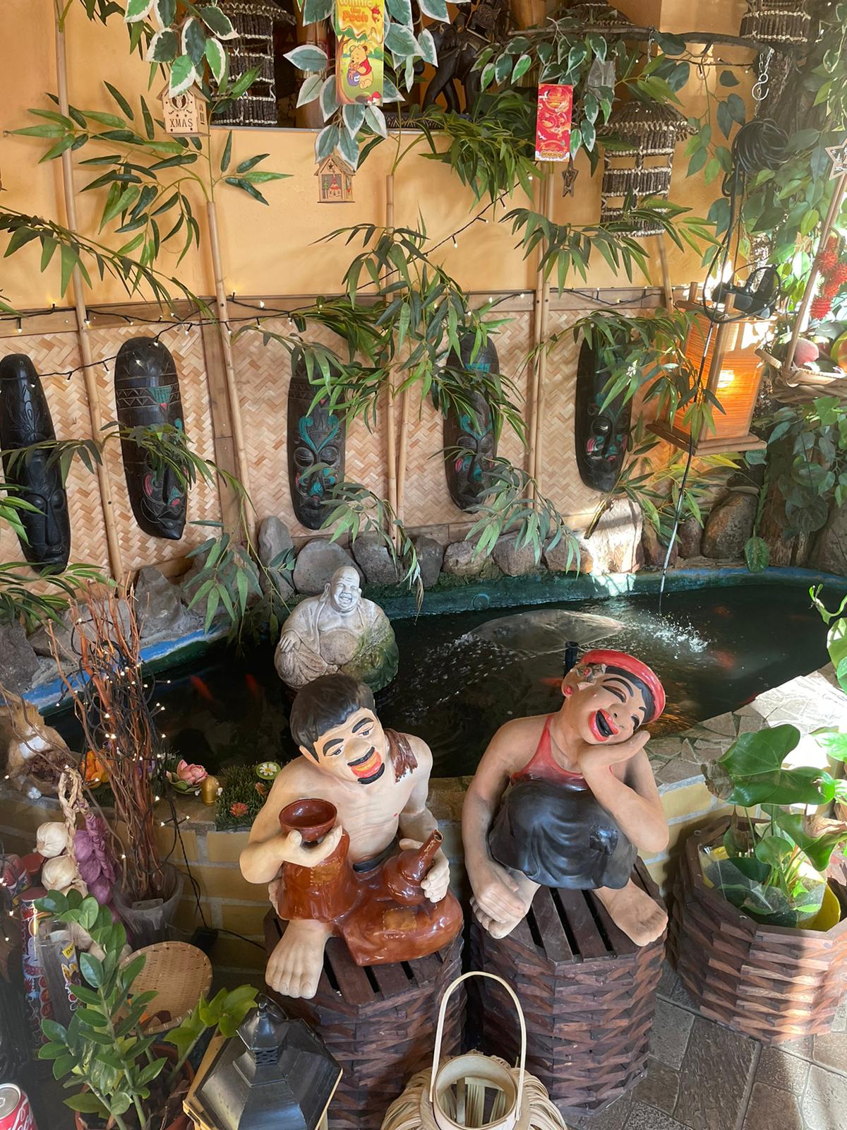Zwei fröhliche, bemalte Tonfiguren neben einem kleinen Teich mit üppiger, grüner Vegetation.